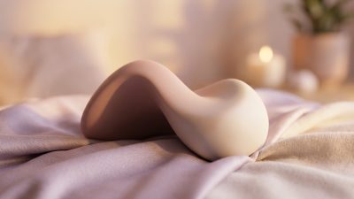 Un objet érotique aux formes organiques et douces posé sur des draps en soie dans une ambiance tamisée.